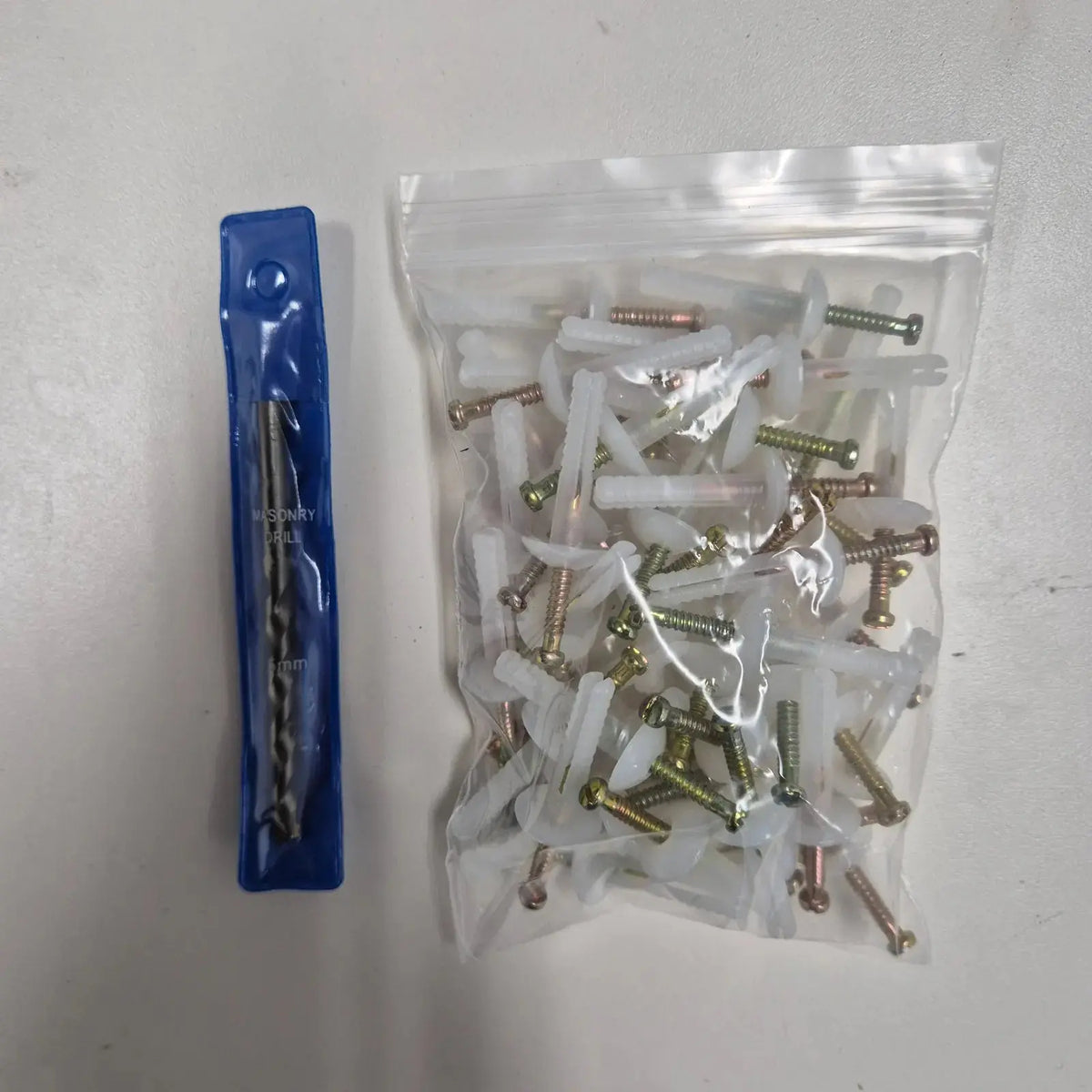 Bag of screws and wall plugs with clips, and a masonry drill bit in blue packaging on a white surface.