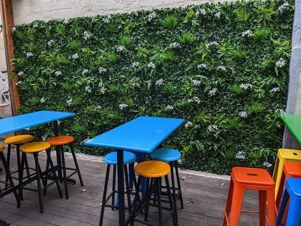 Blue bar tables and colorful stools in front of an artificial green wall with white flowers
