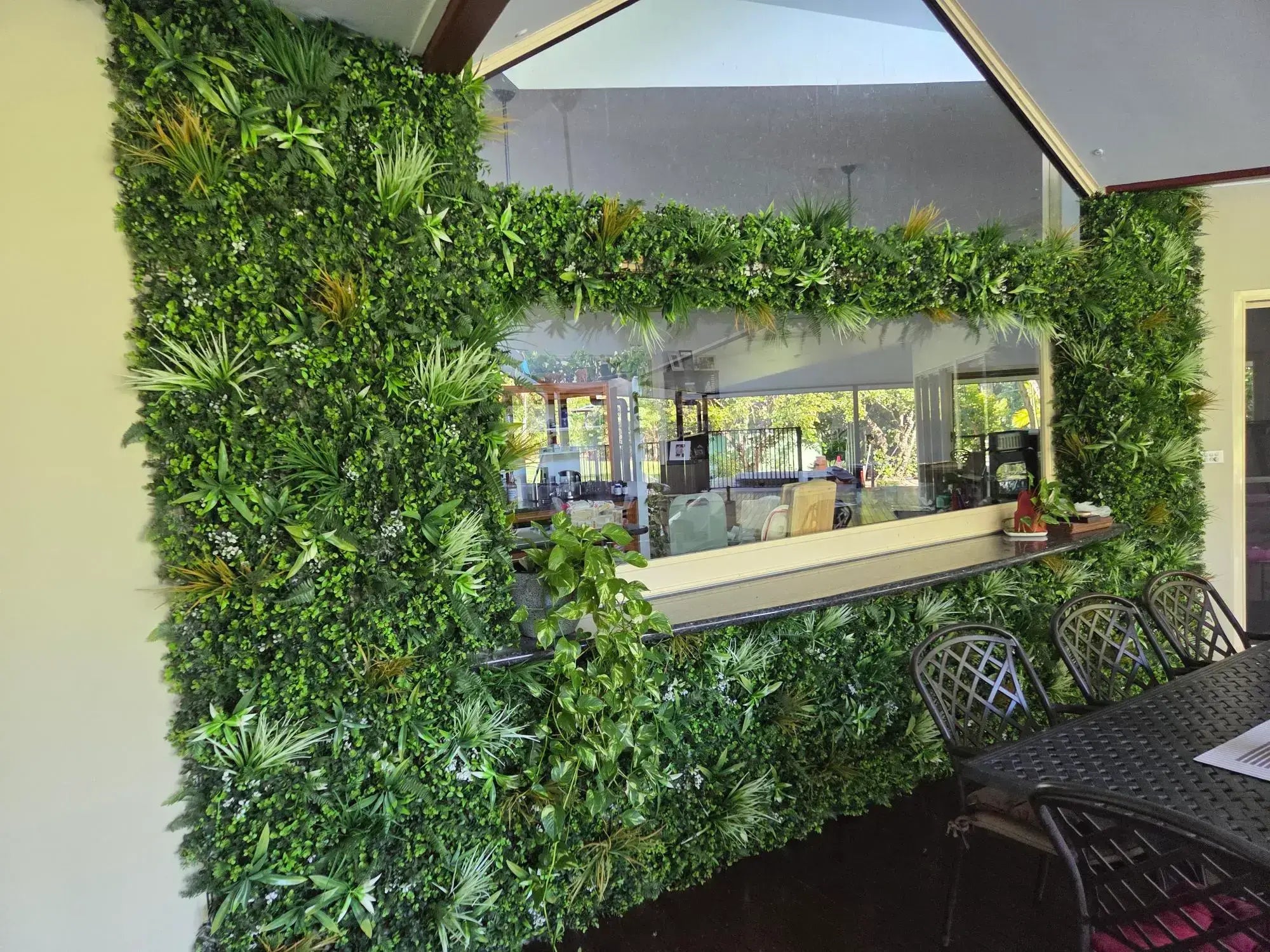 Indoor wall with artificial green plant panels surrounding a serving window and a black metal dining table and chairs.
