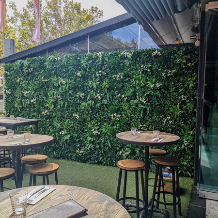Cafe terrace with high tables and stools in front of an artificial green wall.