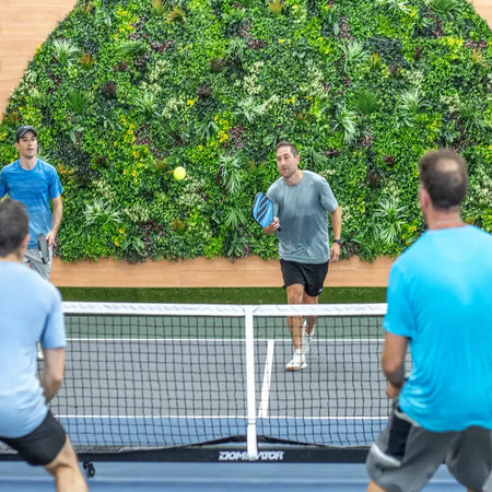 Players on an indoor pickleball court in front of an arched artificial green wall.