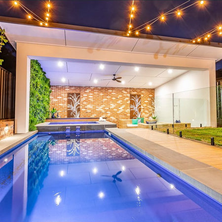 Long lap pool leading to a pavilion with an artificial green wall under string lights.
